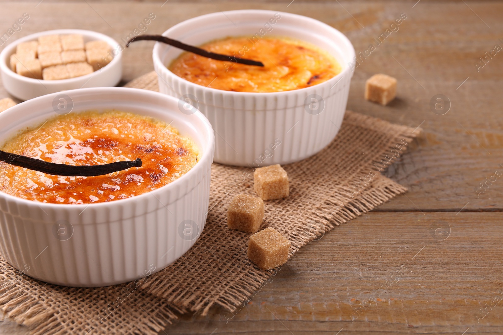 Photo of Delicious creme brulee in bowls, vanilla pods and sugar cubes on wooden table, closeup. Space for text