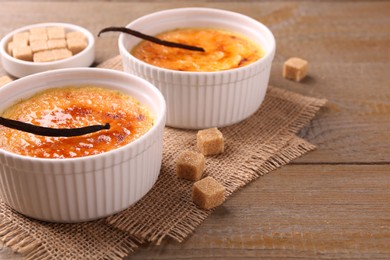 Photo of Delicious creme brulee in bowls, vanilla pods and sugar cubes on wooden table, closeup. Space for text