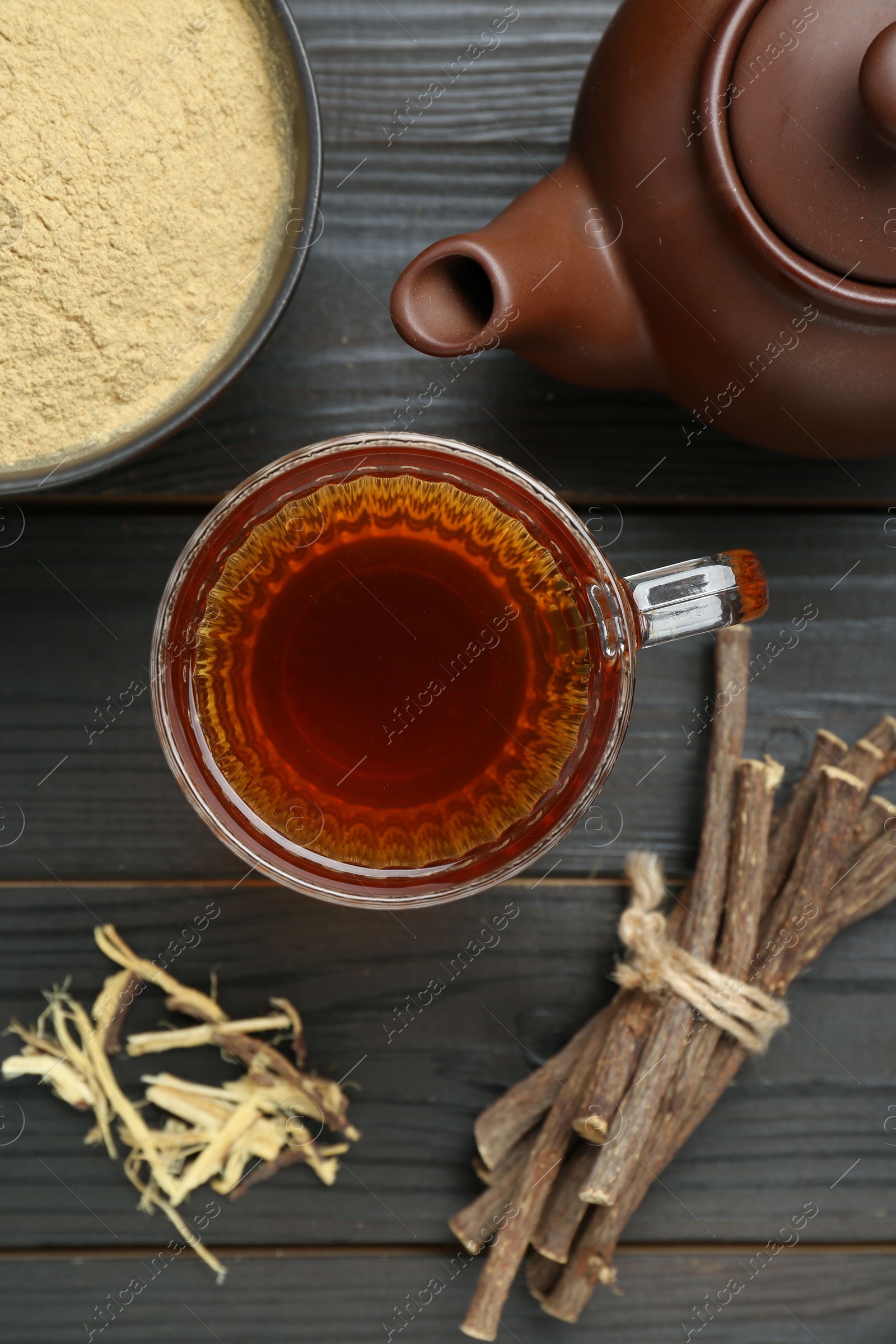 Photo of Aromatic licorice tea in cup, teapot, dried sticks of licorice root and powder on black wooden table, flat lay