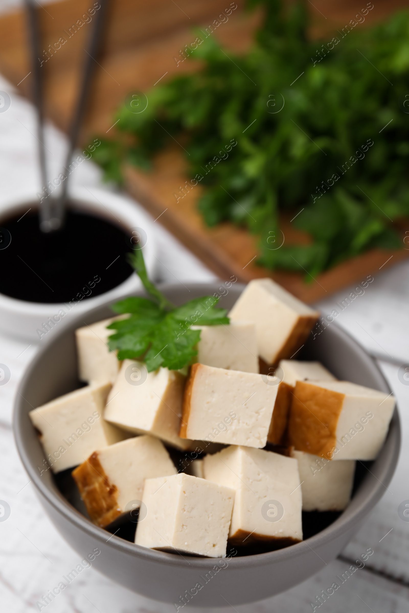 Photo of Bowl of smoked tofu cubes, soy sauce and parsley on white wooden table, closeup