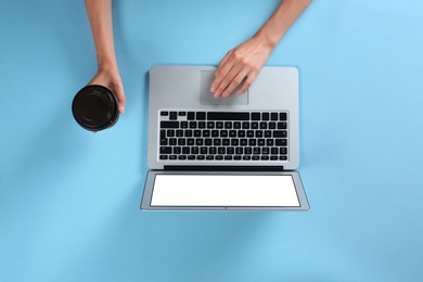 Young woman holding coffee while using laptop on light blue background, top view