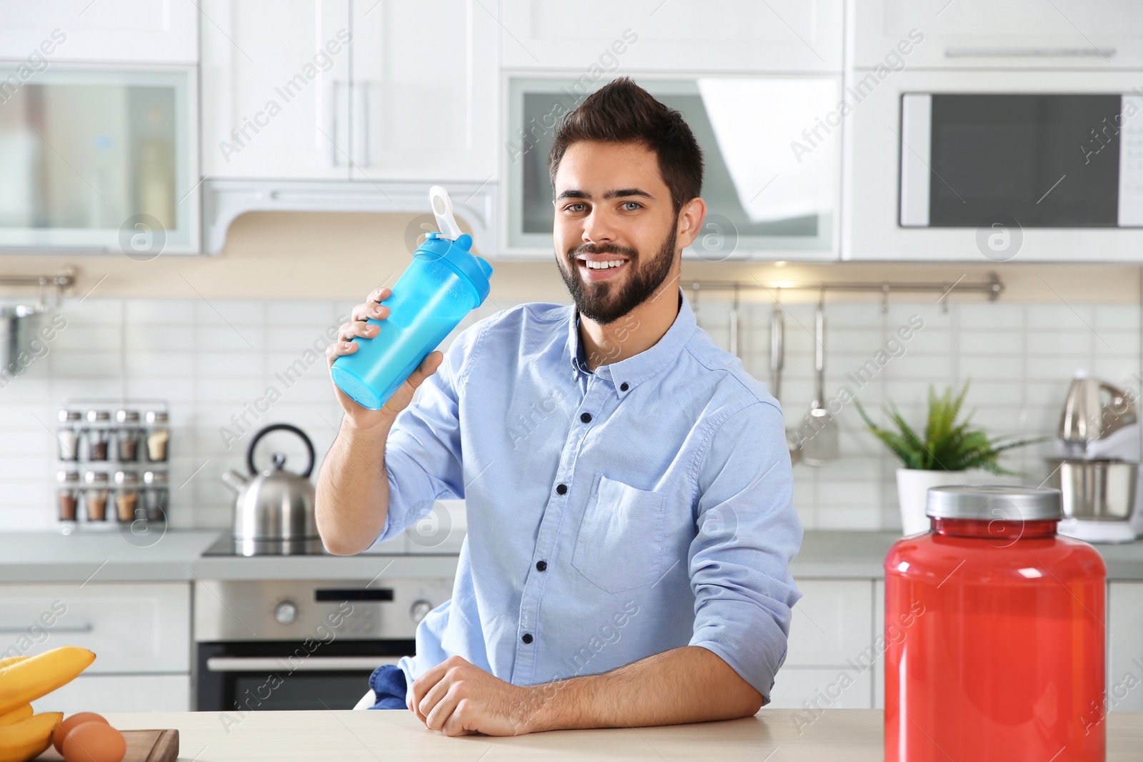 Photo of Young man holding bottle of protein shake at table with ingredients in kitchen