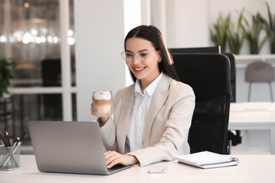 Photo of Happy woman with cup of coffee using modern laptop at white desk in office