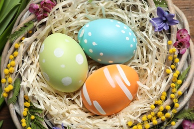 Photo of Wicker nest with painted Easter eggs and flowers on table, closeup