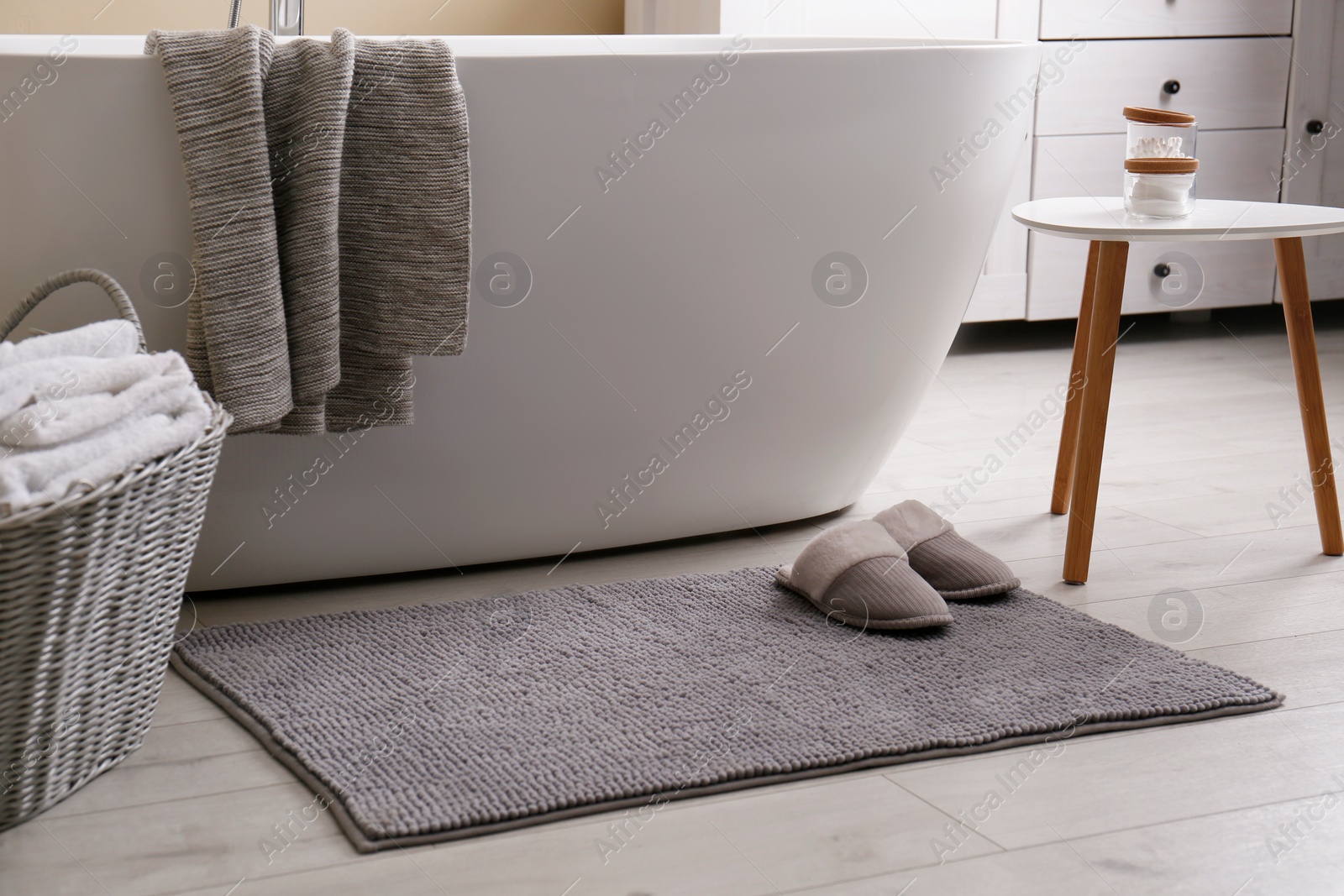 Photo of Soft grey mat with slippers on floor near tub in bathroom. Interior design