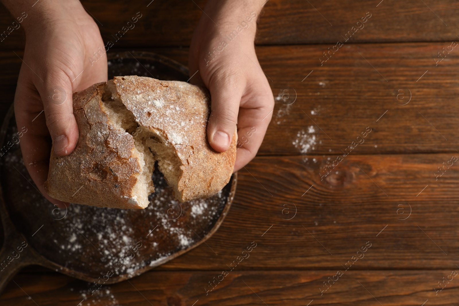 Photo of Man breaking loaf of fresh bread at wooden table, top view. Space for text