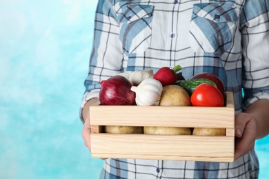 Woman holding wooden crate with vegetables on color background, closeup