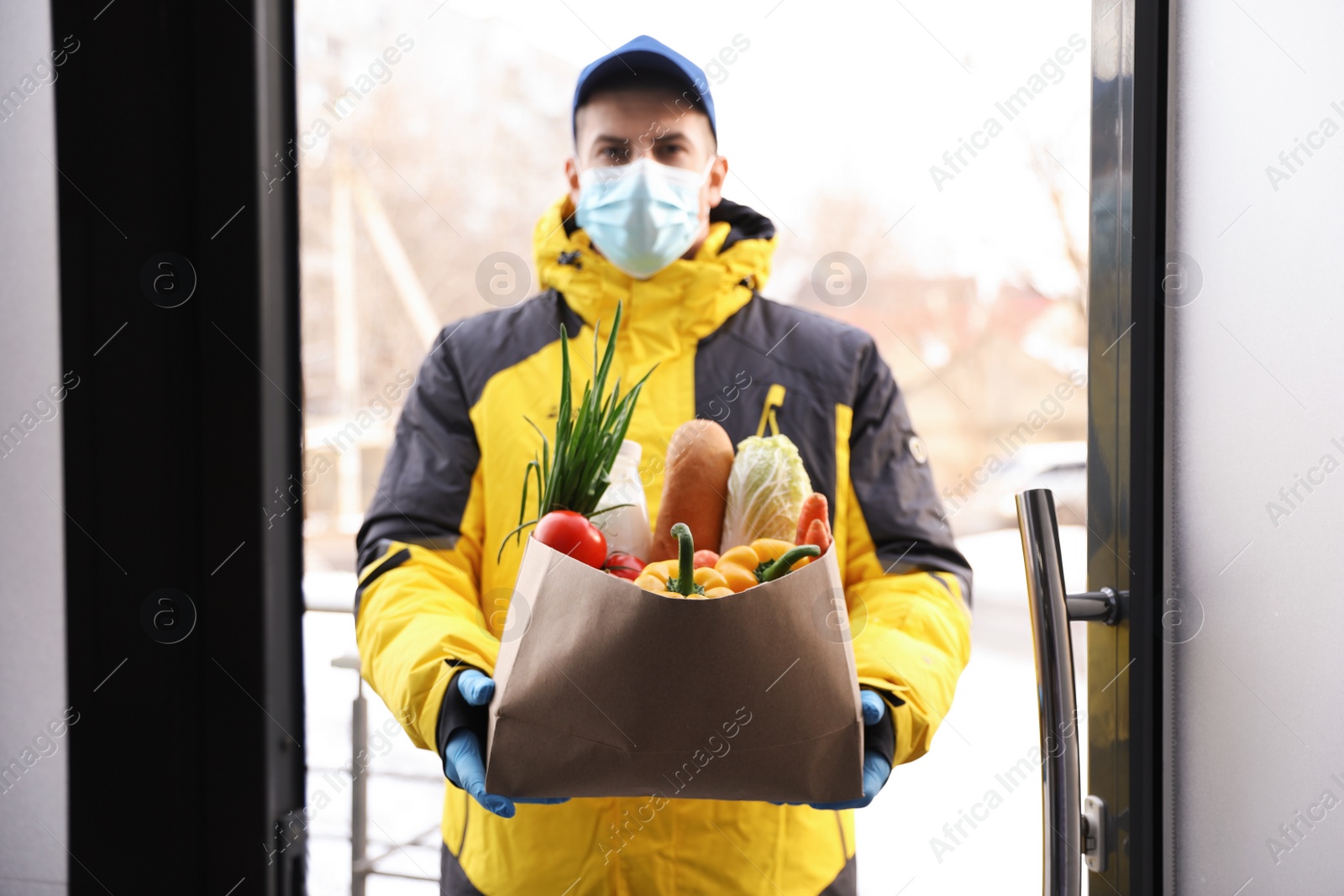 Photo of Courier in medical mask holding paper bag with groceries at doorway. Delivery service during quarantine due to Covid-19 outbreak