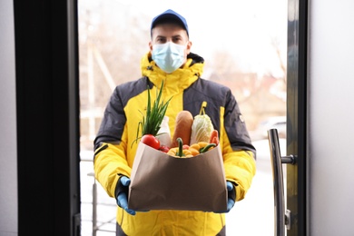 Photo of Courier in medical mask holding paper bag with groceries at doorway. Delivery service during quarantine due to Covid-19 outbreak