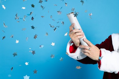 Photo of Man in Santa Claus costume blowing up party popper on light blue background, closeup