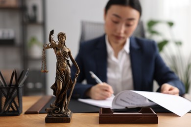 Photo of Notary signing document at table in office, focus on Lady Justice statue