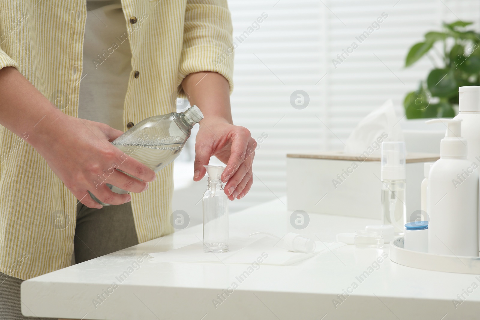 Photo of Woman pouring cosmetic product into plastic bottle at white countertop in bathroom, closeup. Bath accessories