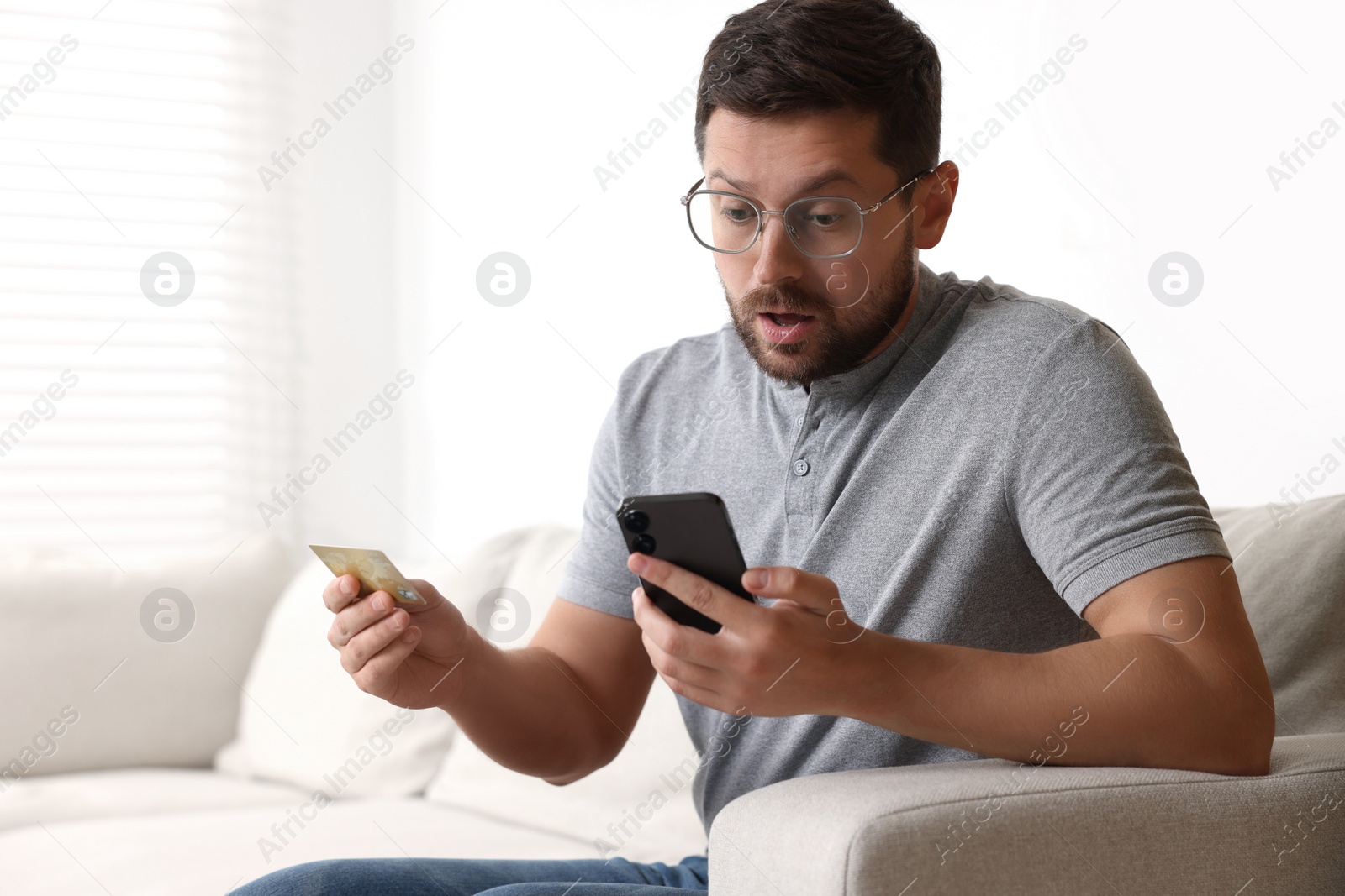 Photo of Shocked man with credit card and smartphone on sofa at home. Be careful - fraud