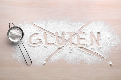 Sifter and crossed out word Gluten written with flour on wooden table, top view