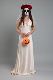 Photo of Young woman in scary bride costume with sugar skull makeup, flower crown and pumpkin bucket on light grey background. Halloween celebration