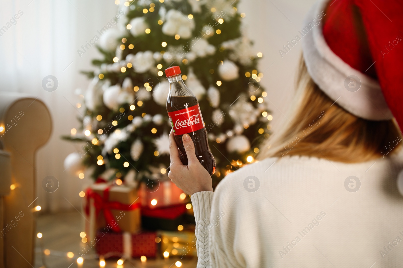 Photo of MYKOLAIV, UKRAINE - January 01, 2021: Woman in Santa hat with bottle of Coca-Cola against blurred Christmas tree at home, back view