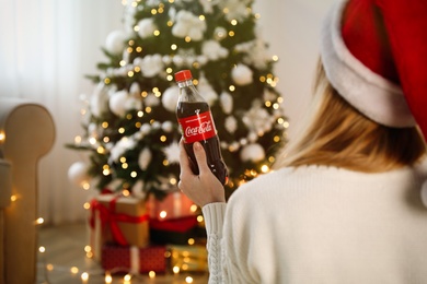 Photo of MYKOLAIV, UKRAINE - January 01, 2021: Woman in Santa hat with bottle of Coca-Cola against blurred Christmas tree at home, back view