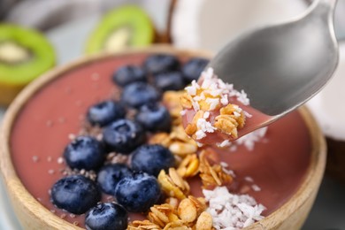 Photo of Spoon of delicious fruit smoothie with fresh blueberries, granola and coconut flakes above bowl, closeup