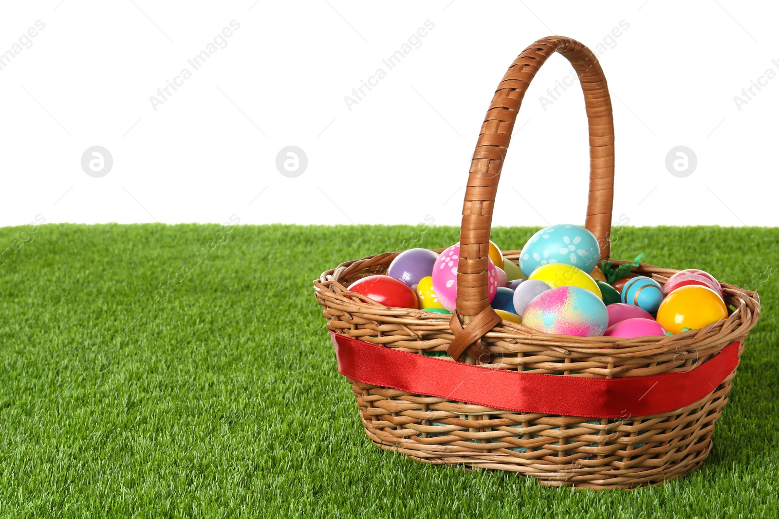 Photo of Wicker basket with Easter eggs on green grass against white background. Space for text