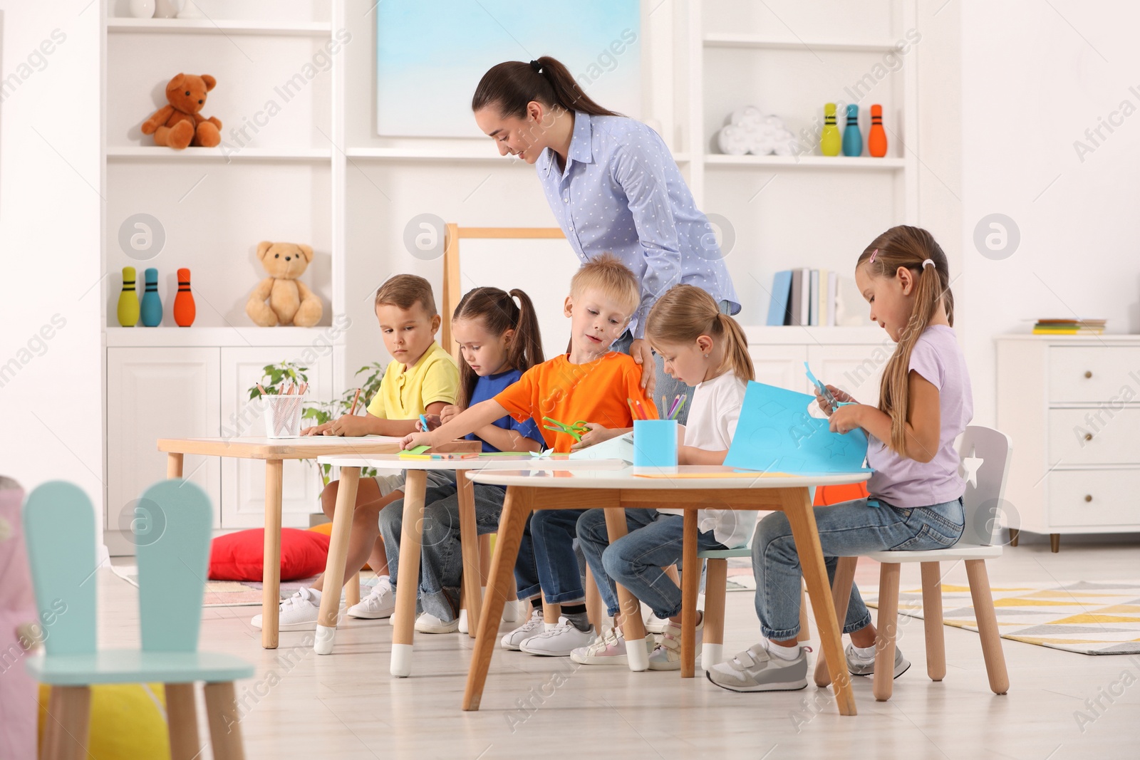 Photo of Nursery teacher with group of cute little children drawing and cutting paper at desks in kindergarten. Playtime activities