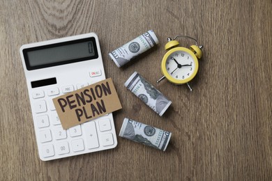 Photo of Card with words Pension Plan, banknotes, calculator and alarm clock on wooden table, flat lay