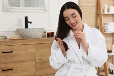 Photo of Happy young woman with bottle applying essential oil onto hair in bathroom