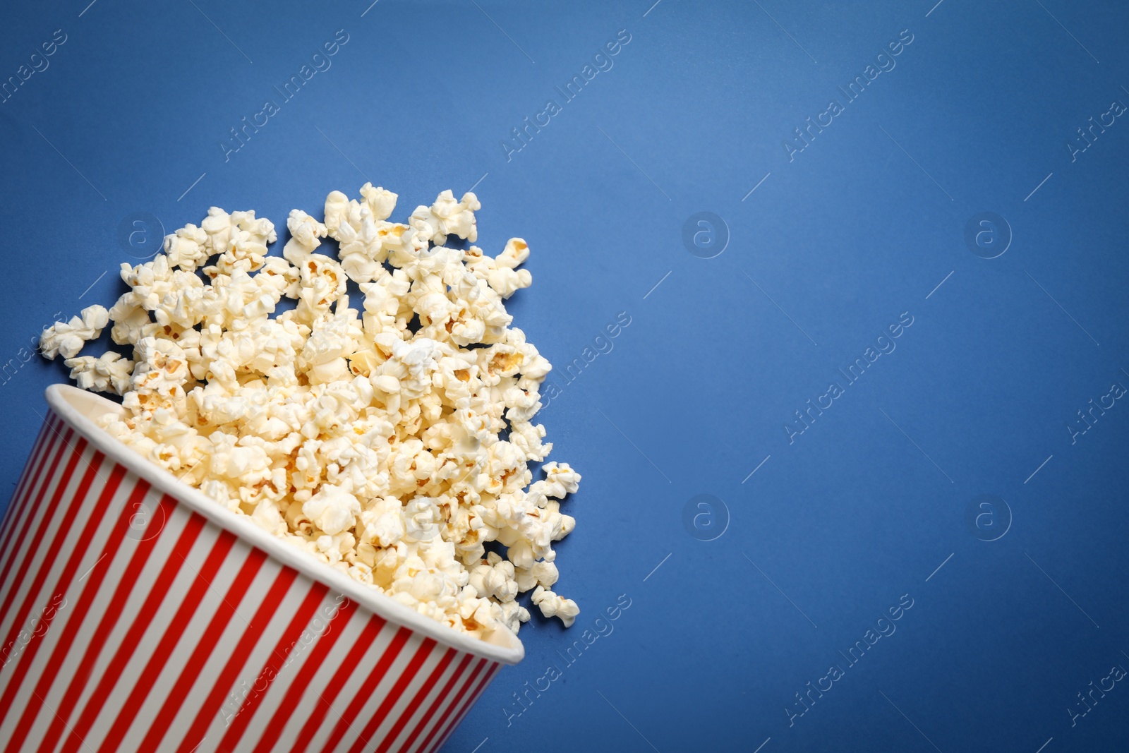 Photo of Overturned paper bucket with delicious popcorn on blue background, top view. Space for text