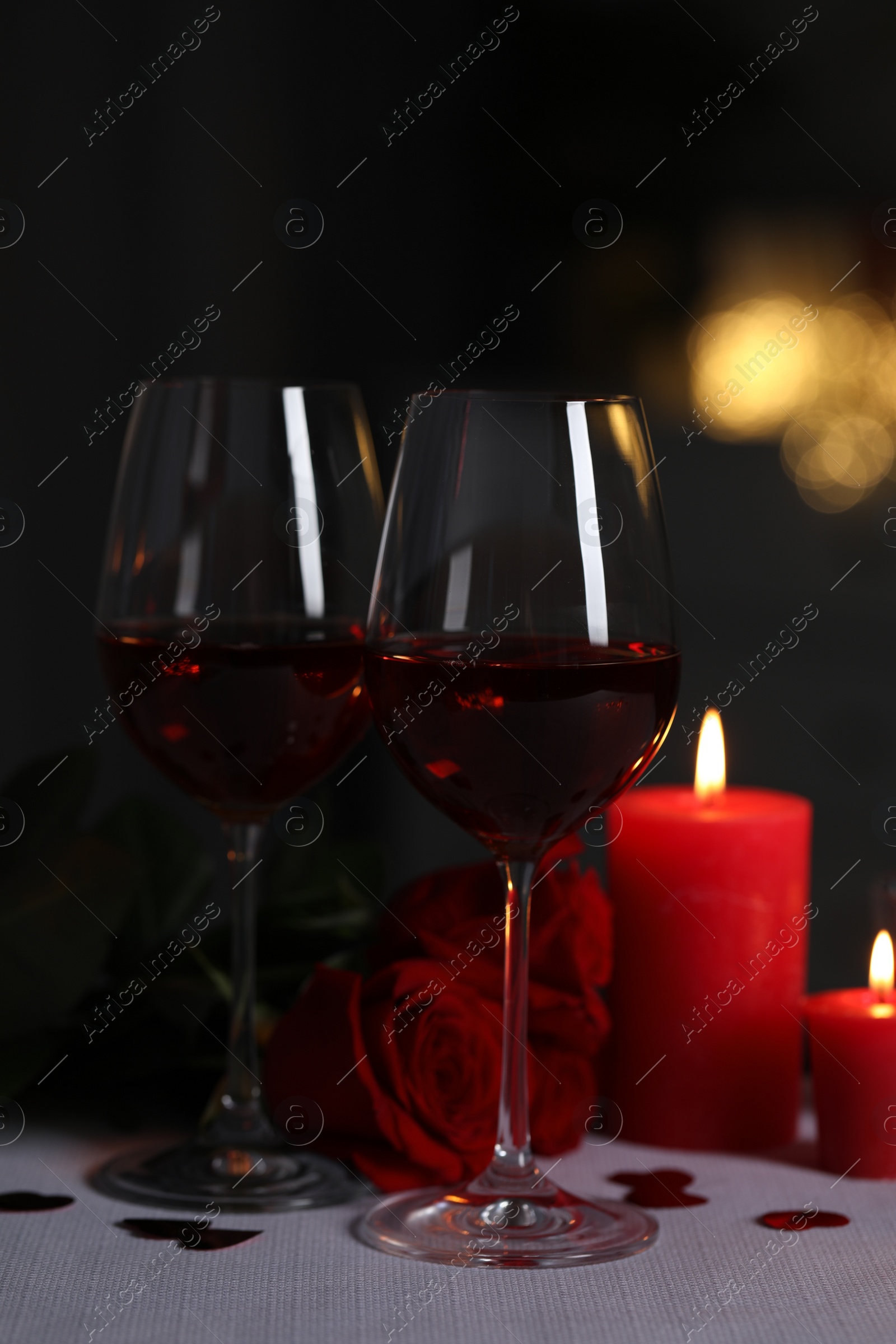 Photo of Glasses of red wine, burning candles and rose flowers on grey table against blurred lights. Romantic atmosphere