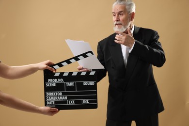 Senior actor performing role while second assistant camera holding clapperboard on beige background, selective focus. Film industry