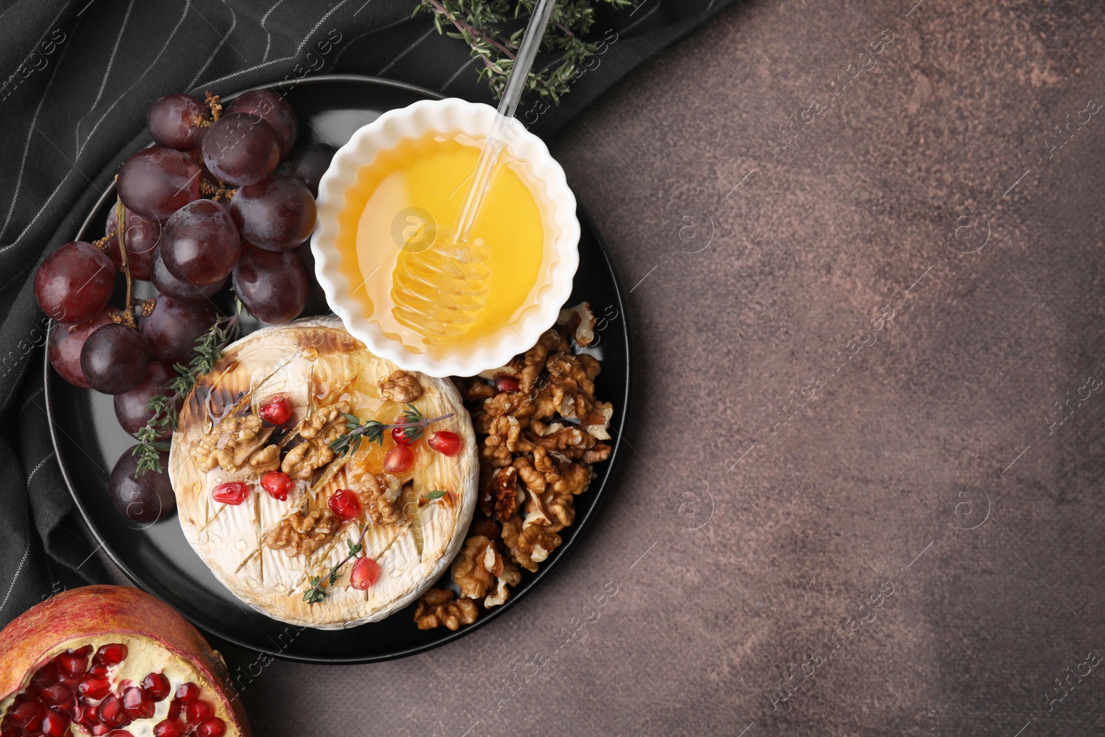 Photo of Plate with tasty baked camembert, honey, grapes, walnuts and pomegranate seeds on brown textured table, top view. Space for text