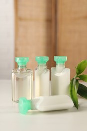 Photo of Mini bottles of cosmetic products and green branch on white table against blurred background