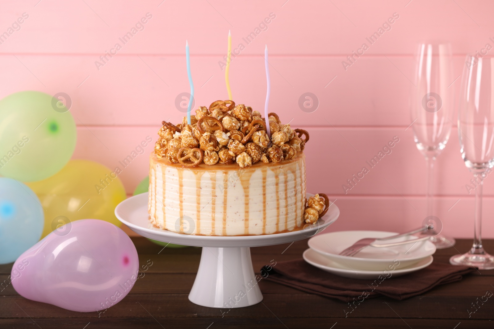 Photo of Caramel drip cake decorated with popcorn and pretzels near balloons and tableware on wooden table