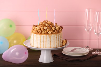 Photo of Caramel drip cake decorated with popcorn and pretzels near balloons and tableware on wooden table