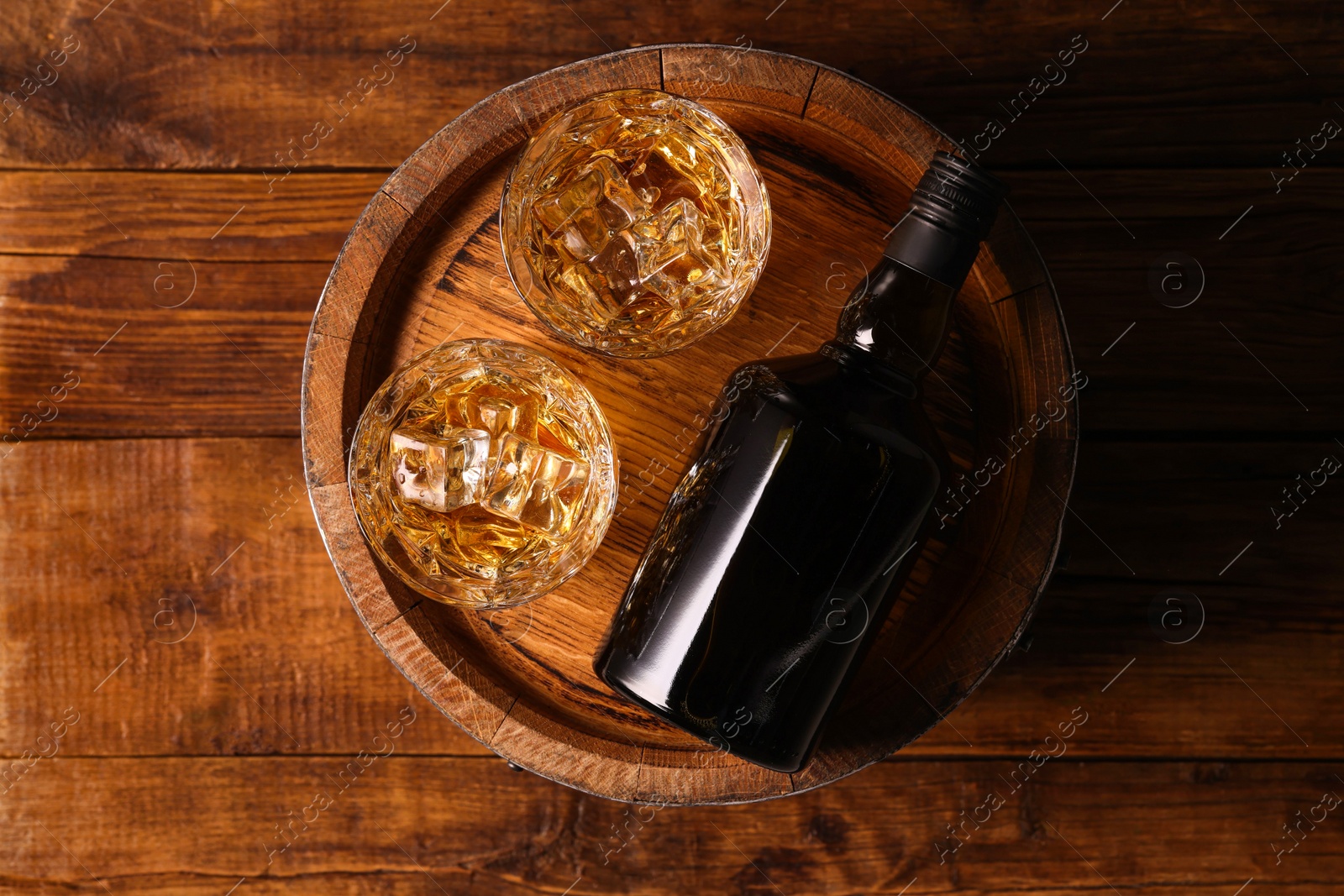 Photo of Whiskey with ice cubes in glasses, bottle and barrel on wooden table, top view