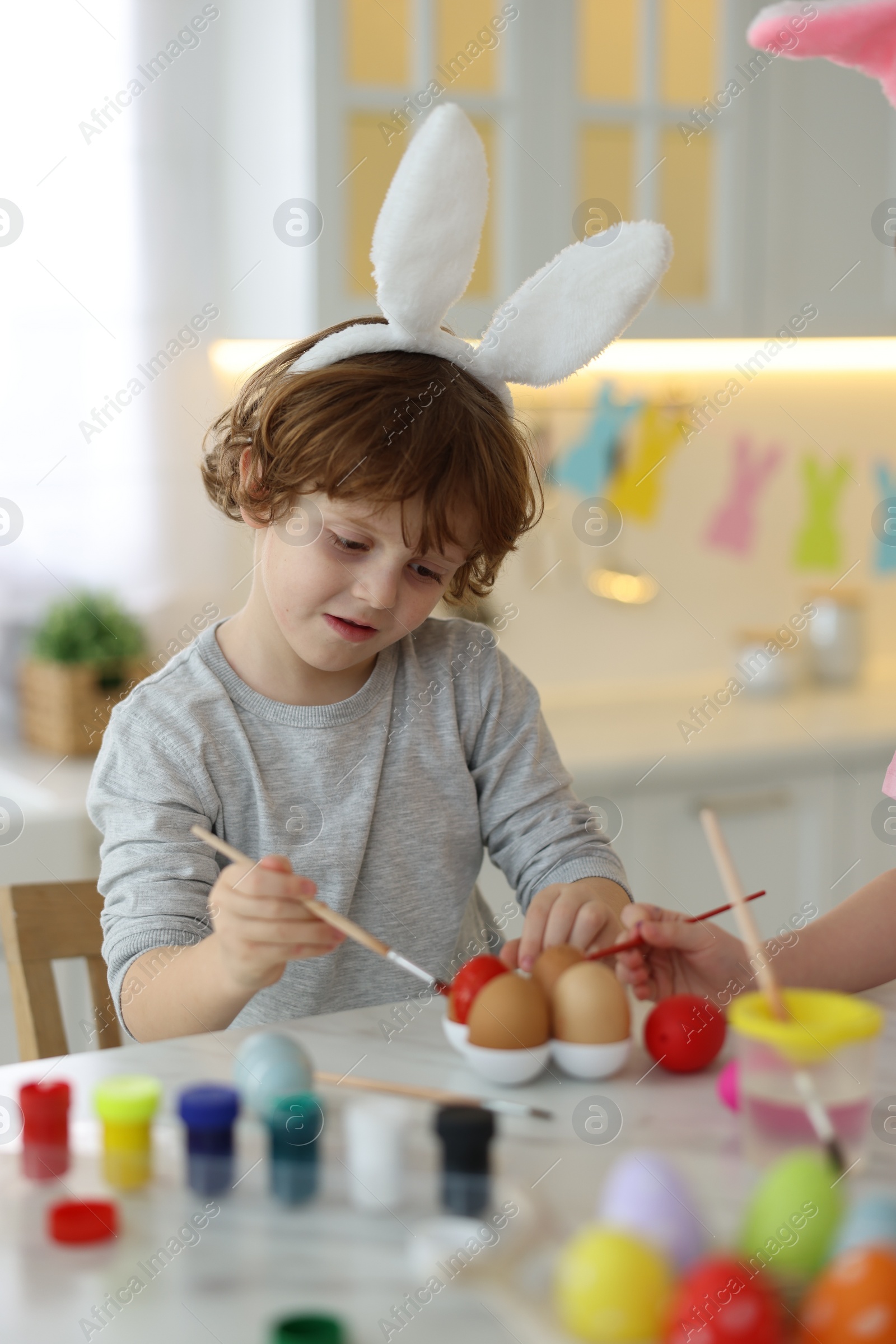 Photo of Easter celebration. Children with bunny ears painting eggs at white marble table in kitchen