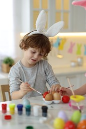 Photo of Easter celebration. Children with bunny ears painting eggs at white marble table in kitchen