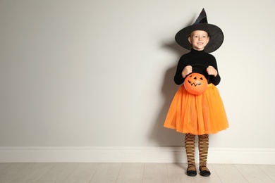 Photo of Cute little girl with pumpkin candy bucket wearing Halloween costume near light wall. Space for text