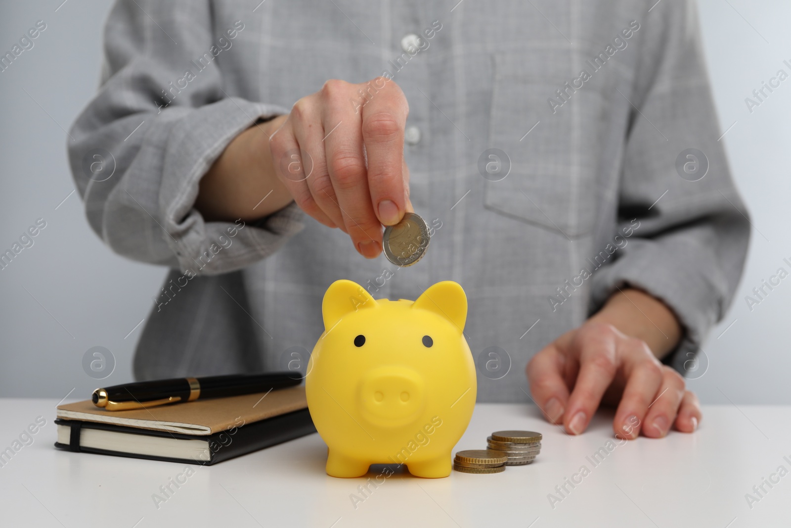 Photo of Woman putting coin into yellow piggy bank at white table, closeup