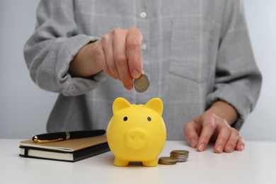 Woman putting coin into yellow piggy bank at white table, closeup