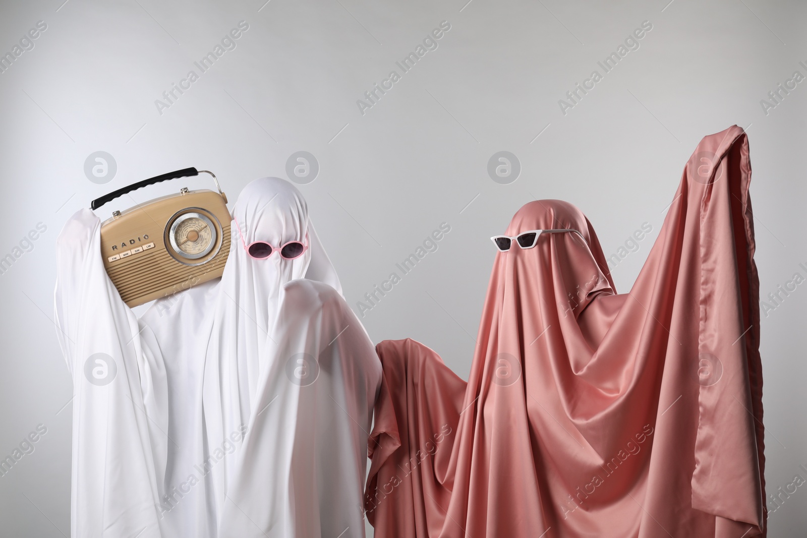 Photo of Glamorous ghosts. Women in color sheets and sunglasses with retro radio receiver on light grey background