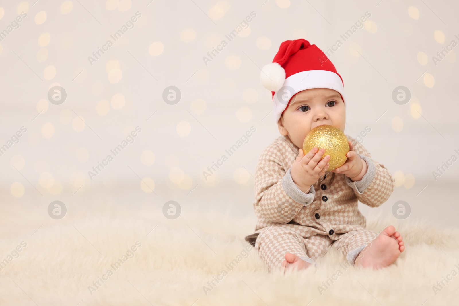 Photo of Cute baby in Santa hat with Christmas bauble on fluffy carpet against blurred festive lights, space for text. Winter holiday