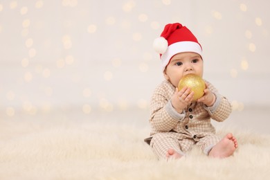 Cute baby in Santa hat with Christmas bauble on fluffy carpet against blurred festive lights, space for text. Winter holiday