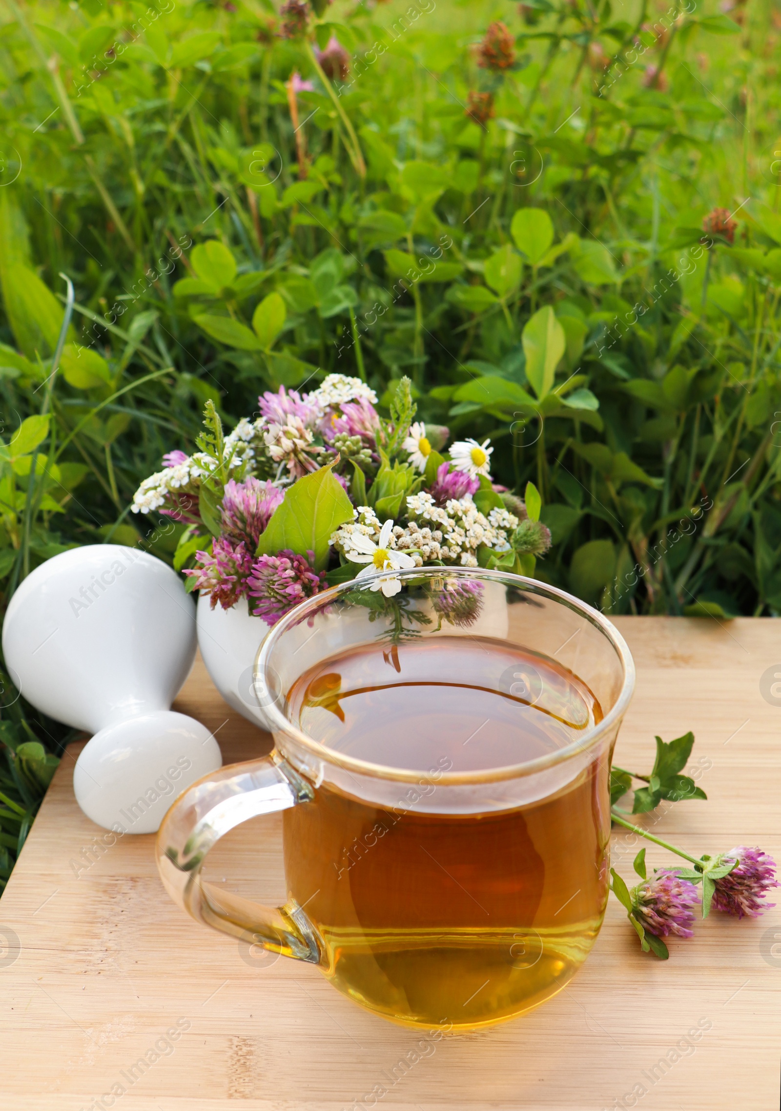 Photo of Cup of aromatic herbal tea, pestle and ceramic mortar with different wildflowers on wooden board in meadow