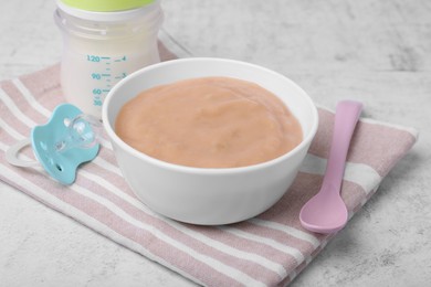 Photo of Bowl with healthy baby food, spoon, pacifier and bottle of milk on white table, closeup