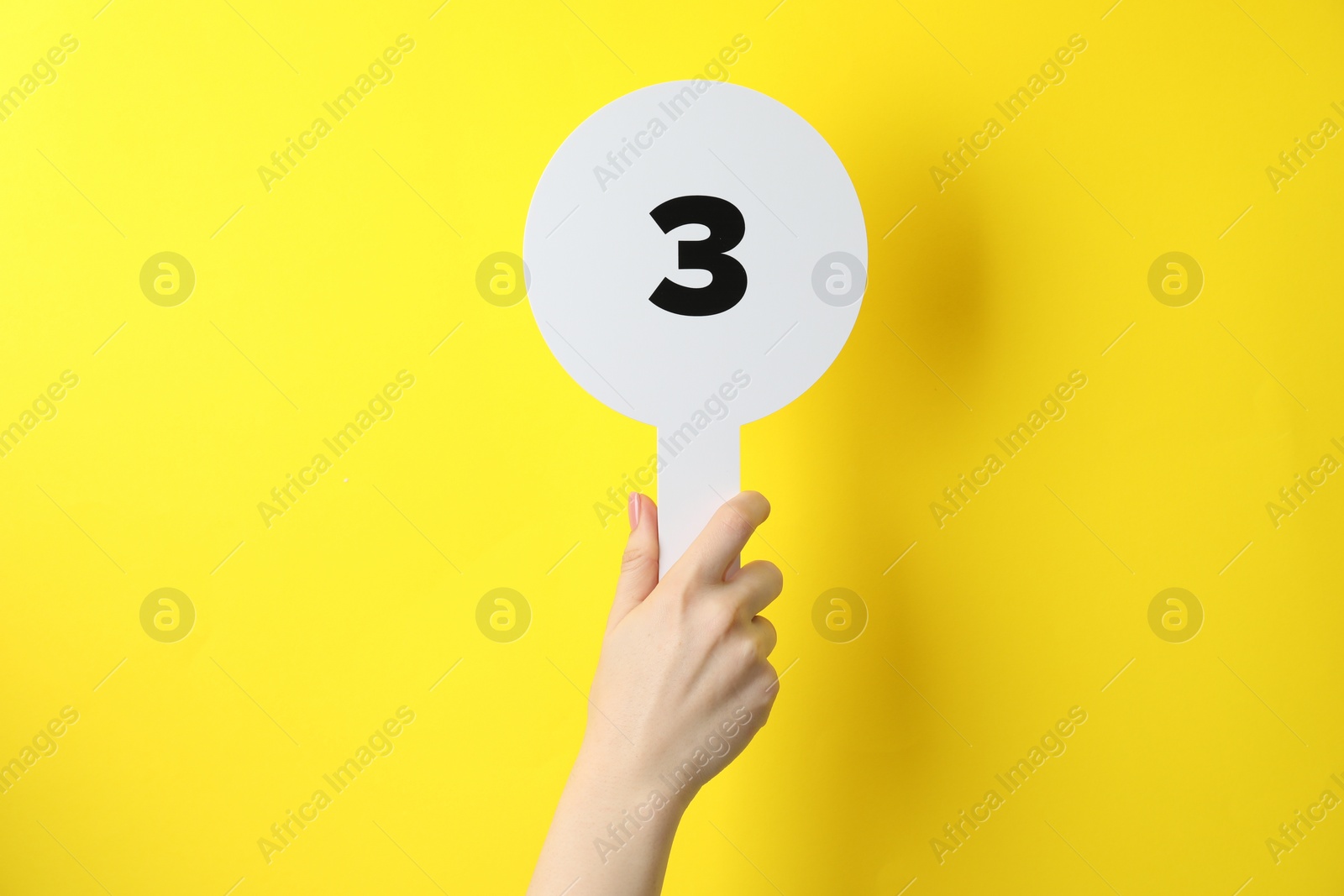 Photo of Woman holding auction paddle with number 3 on yellow background, closeup