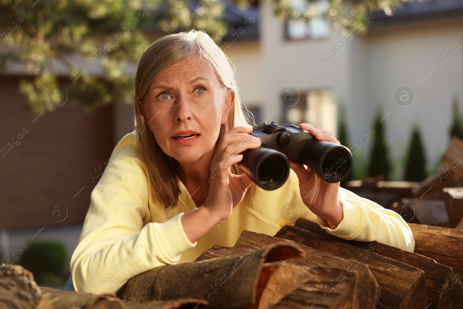 Photo of Concept of private life. Curious senior woman with binoculars spying on neighbours over firewood outdoors