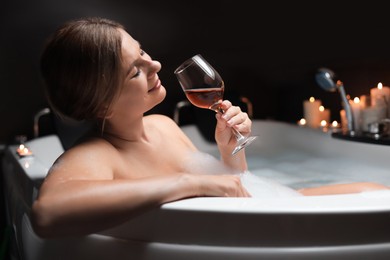 Photo of Beautiful woman drinking wine while taking bubble bath indoors. Romantic atmosphere