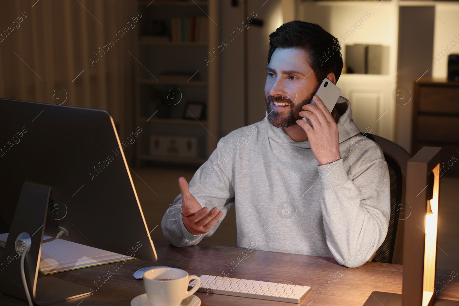 Photo of Home workplace. Happy man talking on smartphone while working with computer at wooden desk in room at night