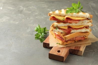 Stack of tasty sandwiches with ham, parsley and melted cheese on grey textured table, closeup. Space for text