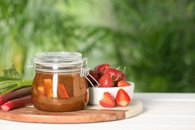 Photo of Jar of tasty rhubarb jam, fresh stems and strawberries on white wooden table against blurred background. Space for text